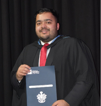 Umang Agrawal wearing a black graduation gown with a red tie, holding a certificate standing in front of a black curtain