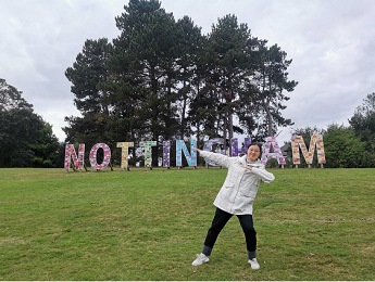 Xiaojie Cheng wearing a white coat and blue jeans standing on grass and pointing at large letters spelling NOTTINGHAM