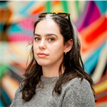 Photo of Amy Miller. Amy is stood in front of a multicoloured background and has long dark hair. Amy has a pair on sunglasses on the top of her head and is wearing a grey jumper.