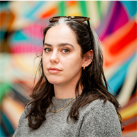 Photo of Amy Miller. Amy is stood in front of a multicoloured background and has long dark hair. Amy has a pair on sunglasses on the top of her head and is wearing a grey jumper.