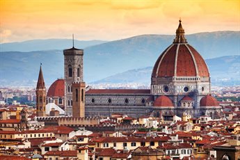 Photo of the cathedral in Florence, Italy at sunset. Buildings can be seen in the foreground and mountains are visible in the background. The sky is hues of orange and yellow.