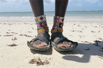 Photo of a pair of feet standing on a white sand beach wearing colourful cultural ankle braclets.