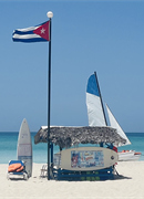 Photo of a white sandy beach with blue sky and a blue ocean. A beach hut with sailing boats and windsurfers are on the sand beside the hut.