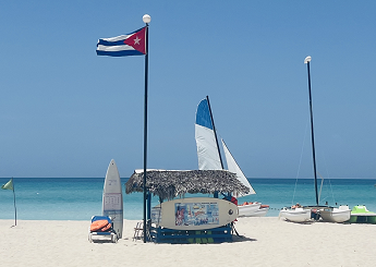 Photo of a white sandy beach with blue sky and a blue ocean. A beach hut with sailing boats and windsurfers are on the sand beside the hut.