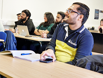 students in classroom sitting at tables with laptops and books facing the front