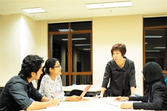 A team sit and stand around a boardroom table covered with papers and laptops. A woman stands talking to the team at the head of the table whilst the other three team members, two women and a man, listen.
