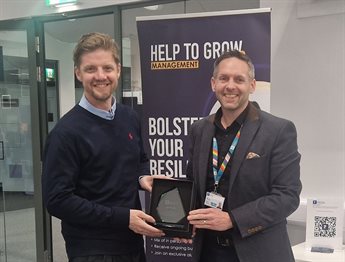 Antony Rhodes receives an award from Executive Education staff member Nick Proverbs. They stand together in front of a Help to Grow:Management programme banner, holding the award between them and both smiling at the camera.