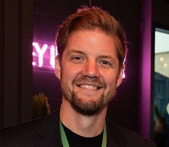 Profile photo of Antony Rhodes. Antony is shown in a darkened room, backlit by a purple neon light. He smiles at the camera.