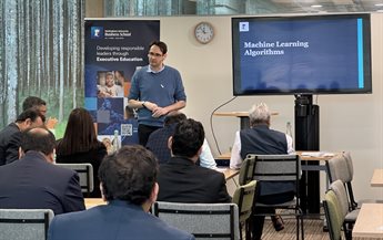 Participants from the NIBM programme listen to a speaker. They sit around tables facing the speaker who is standing next to presentation screen which reads 'Machine Learning Algorithms'