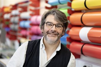 Tom Hignett, a Graphic Design Technician/ Demonstrator from the University of Chester, stands in a design studio. He is dressed in smart-casual clothes and smiles at the camera. Behind him, rolls of coloured vinyl are mounted on the wall..