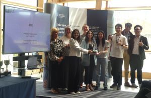 MSc Company Consultancy Competition presentation. Team for Jaguar Land Rover, winners standing in front of a large banner holding a silver trophy: mentor Professor Andrew Callaghan from Business School, Business partner Lee Cooper-Blair, students Gunjan K