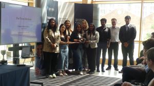 MSc Company Consultancy Competition presentation. Team for The Treat Kitchen, winners standing in front of a large banner holding a silver trophy: mentor Professor Professor Keith Burgess from Business School, Business partners Jess Barnett and Lydia McDo