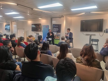 MSc Marketing students sitting down facing the front of a room at Ipsos in London with 2 people sitting on stools, Colin Strong and another speaker