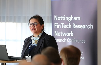 Meryem Duygun wearing a dark blue jacket and blue neck scark standing sideways in front of a dark blue banner with white letters - Nottingham FinTech Research Network