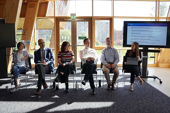 People sitting in a row presenting in front of large windows and a screen to the right at the OMIS Workshop May 2025