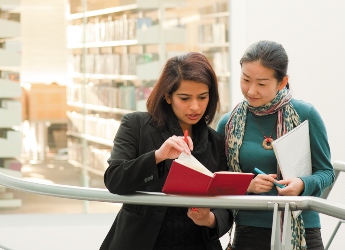 Two female students in library looking at a red book