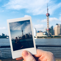A hand holds a polaroid shot of a tower building in china, directly in front of the real thing