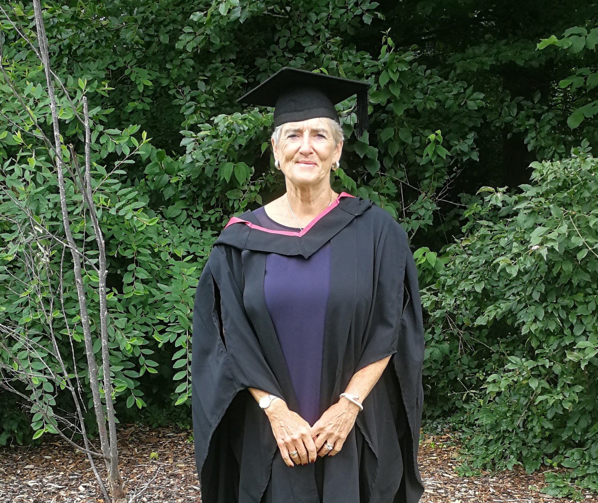 Fiona on graduation day, wearing a black motarboard hat and gown, holding her degree certificate
