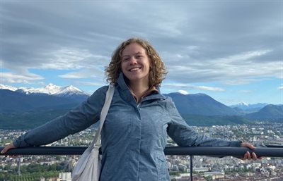 Photo of Amy Groves smiling while leaning on a railing, with a scenic mountain and cityscape in the background