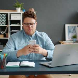 A man at a desk, using his phone with a laptop and pad on his desk