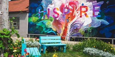 A colorful outdoor mural with the word &amp;quot;INSPIRE&amp;quot; painted in large, vibrant letters, set against an abstract background with floral and butterfly designs. In front of the mural is a small garden with plants, flowers, a bright blue bench, and a chair. A bui
