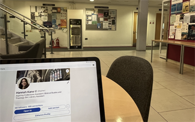 Photo of student Linkedin profile on an open laptop in the foreground. In the photo background, stairs and water dispenser can be seen.