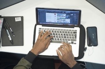 Person typing on a laptop with design software open, surrounded by a notebook, pens, and a smartphone with earphones.