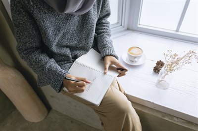Photo of a lady sat on the windowsill writing in a notepad. Beside her is a cup of coffee and some decorations