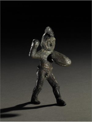 Ancient bronze statue of a standing Spartan warrior with helmet and shield. One upraised hand is empty but used to contained a spear. Roughly formed.