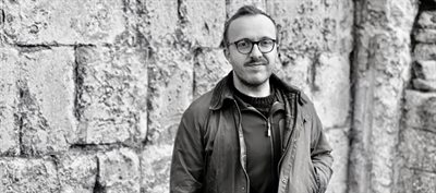 Black and white photo of a young white man wearing an anorak with short hair and glasses. He is standing in front of an old wall constructed from large stone blocks.