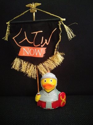 A yellow rubber duck in Roman armour, holding a Latin Now banner against a black background.