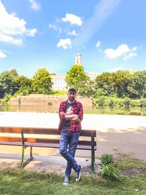 A bearded man (Fabio Saccoccio) wearing jeans, a Ghostbusters t-shirt beneath a red checked shirt and a cap leans casually against a bench with the University of Nottingham Trent Building behind him on a beautiful sunny day.
