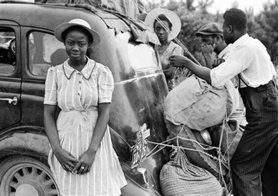 Black and white historical photograph of a young Black woman in a sun hat and dress stood beside a car. Other family members converse in the background.