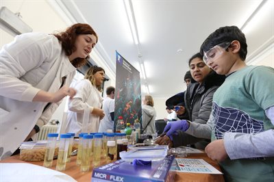 Female scientist in white lab coat talking to a child the other side of a table as he undertakes a table top experiment, while wearing safety googles