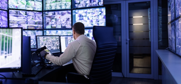 Man sitting in front of multiple computer screens displaying surveillance video
