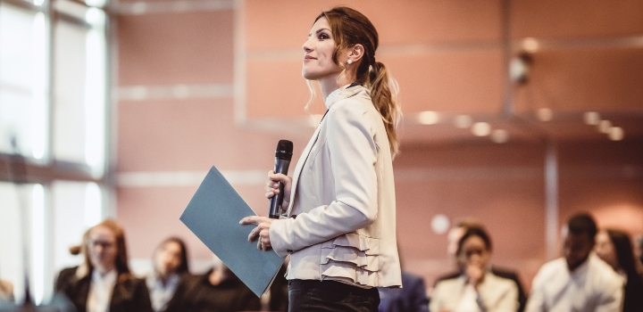 Woman holding a microphone looking at screen in front of an audience