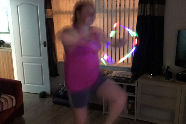 A soft focus image of a woman in shorts and a bright pink t-shirt taking part in a live lockdown areobics to rave music class using a disco ball and bright glowsticks!