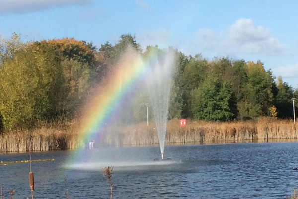 A photo of a blue lake with a white fountain, taken on a bright sunny day just after a rain shower where the fountain and light have combined to form a rainbow under the bright blue sky.