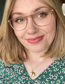 A close-up headshot of Lizzie Alblas, smiling and wearing a green patterned dress and clear glasses