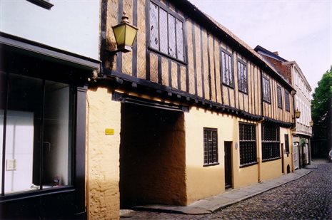 Modern photo of an old house on a street with cream coloured walls and dark timber framing in vertical rows along the outside of the upper floor.