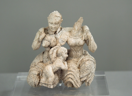 An ivory figure of two women and a child, the woman on the right is missing her head. The child leans across their laps.