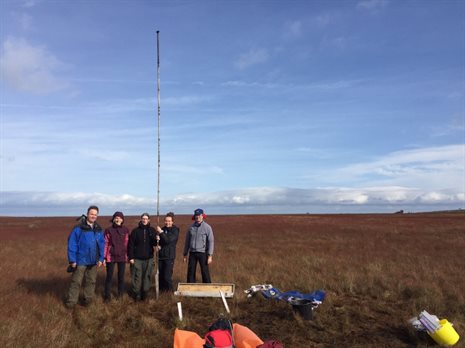 Five people, three women and two men, in coats stand facing the camera holding a large pole with other equipment and objects scattered around them and a flat, grassy plain below blue skies behind.