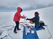 Two people in thick coats working surrounded by equipment on a snowy landscape.
