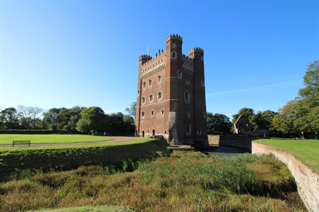 Tall crenellated tower in red brick surrounded by trees and a large field on one side with a clear blue sky behind