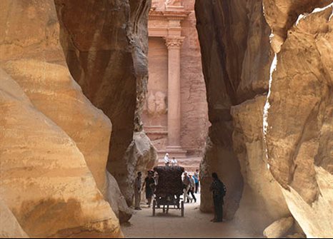 Image of cart driving away from the camera, along a narrow dirt road between two pale stone cliffs that frame the image on either side. In the background are crowds of people in front of the city of Petra.