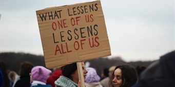 A person in a crowd holding up a sign which reads "What lessens one of us lessens all of us".