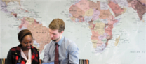 A smartly dressed man in a tie in discussion with an African lady, in front of a large wall print map of the globe
