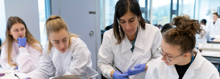 An academic and three students wearing lab coats and looking down at a desk, which is out of shot.
