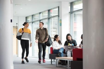 A female student and a male student in conversation walking past three seated female students.
