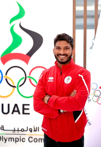 Dr Vishnu smiling for the camera with his arms folded, standing next to a sign for the UAE Olympic Committee